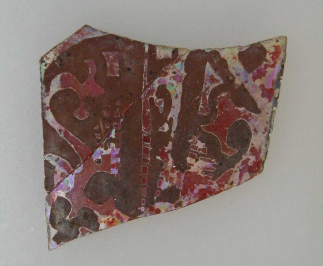 1 fragment af rude med afnappede  kanter af blegr�dt glas med bemalet stiliseret plantemotiv i brunlig farve. Hidr�rer fra rude af langstrakt form let afsmalnende, men kun den ene halvdel med den bredde konkavt indsvungne endeside er bevaret. St�rste m�l: ca. 4,0 x 3,6 cm.