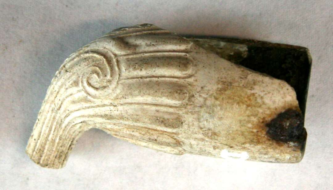 1 fragmenteret kridtpibehoved af konisk let hv�lvet form uden h�l og med delvis afbrudt munding. Den bevarede stump af piber�ret er dekoreret med t�tte fine ribber, hvoraf nogle afsluttes i en oprullet spiral nederst p� hver side af hovedet, andre danner en spinkel indramning om den krans af vulsttunger, der l�ber halvvejs op p� siden af hovedet. S�vel hovedets som r�rets ydre og indadvendende midterlinie er markeret ved en skr� rifling, der forts�tter op p� vulsttungerne. P� hovedets indadvendte side findes umiddelbart over den riflede tunges afslutning et indstemplet cirkul�rt v�rkstedsm�rke hvori en bugtet slang i oph�jet relief.