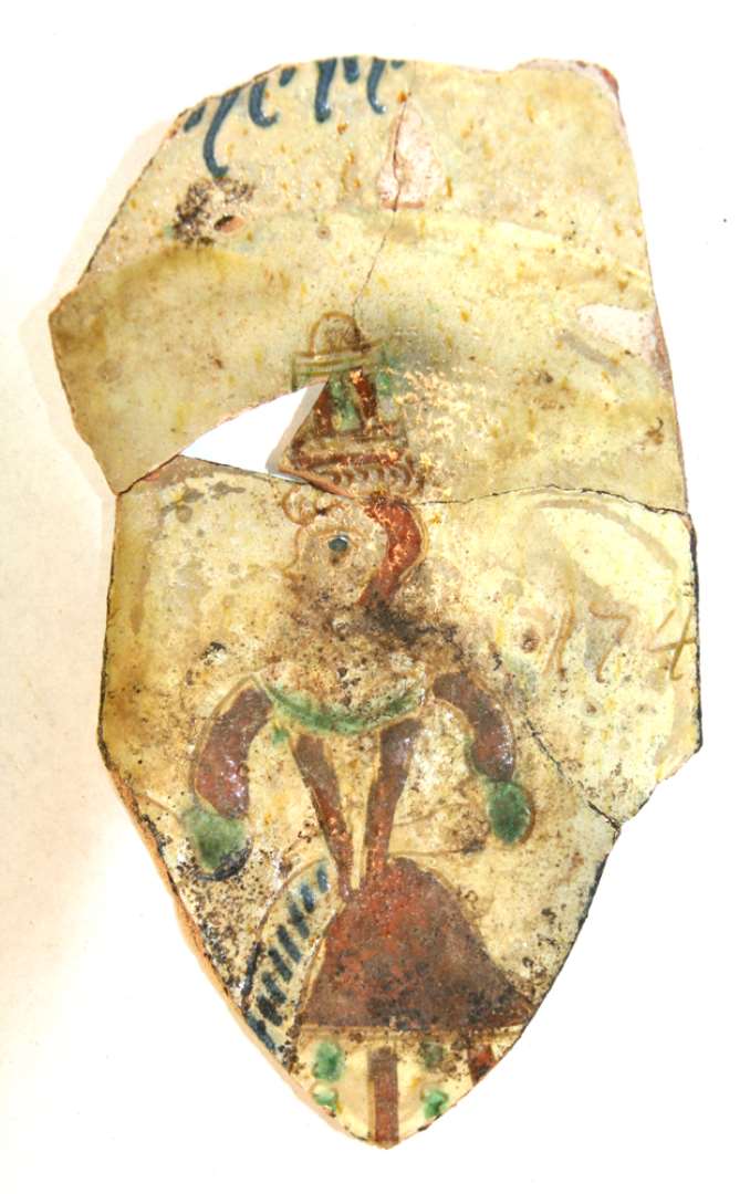 Bund/sidefragment samlet af tre sk�r fra fad af gulligt lergods med pibelersbegitning og polykrom dekoration. P� fadets bund en delvis bevaret, stiliseret kvindefigur i folderig kjole med bred brystudsk�ring, smalt, tilspidset liv og albuelange �rmer i brunt, gr�nt og bl�t, p� hovedet: h�j kunstf�rdig h�ropbygning i brunt og gr�nt. Til h�jre for kvindens skulder: 174(?) med svagt indridsede tal udfyldt af glasurmasse. P� fadets rand spor af en oph�jet indskrift i bl�t baggrundsglasuren er overalt let sp�ttet, grumsetgullig. Bunddiameter 9,5 cm.