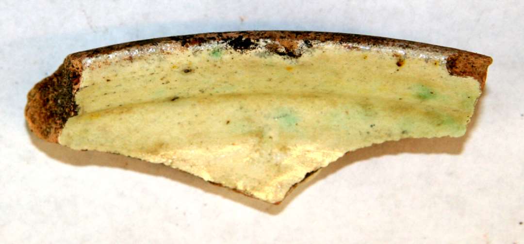 1 randsk�r fra fad af gulligt lergods med kraftig ydre mundingskrave. Indersiden er d�kket af pibelersbegitning, hvorover klar, svagt gr�nligsp�ttet glasur.