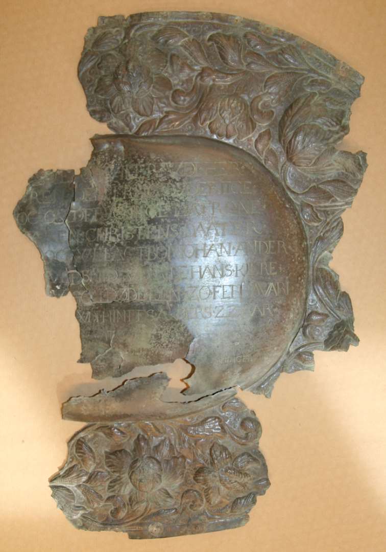 Brudstykke af kisteplade af messing. �Oval form. Ovalt buklet midtskjold, omgivet af blomsterramme, udf�rt som foreg�ende.�P� pladen indgraveret: 'HER.HVILER.VDI.HER./ REN.DEN.ERLING.DYDERIIGE/.OC.GVDFRYGTIGE.MATRONE./ (M)AREN.CHRISTENS.DAATER./ OC.VELACTBOR.IOHAN.ANDER./ ...(G)VLDSMID.WDI.RIBE.HANS.KIERE./ ...RV(.)SOM.D�DE.DEN.20.FEBRAVARI./...WDI .HINDES.ALERS.22.AAR./APOC.14:/ ...�DE.SOM.D�.HEREFTER.I.HERREN:./ ...AF.DIERIS.ARBEYD.OC.D(IER)IS.GIERNINGER:./ �...R.DE(M)? EFTER:.'