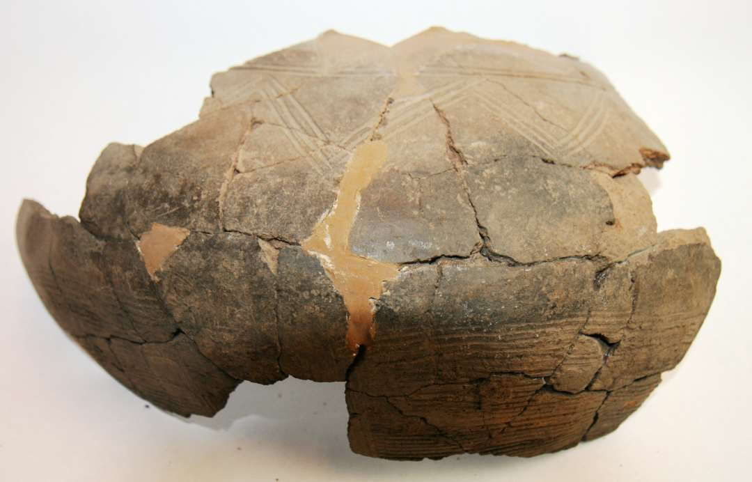 Del af rand og bug til finere Krukke - Rand tynd, lige afsk�ret, p� udsvajet hals der i kraftig bue g�r over i bug. Overgang til skulder markeret med bred fure, derunder to overl�bende streger og derunder h�ngene vinkler, dannet af tre (3) streger. Nederste bevarede del af bug med kamstreger.  M�rkbrun glittet gods. 